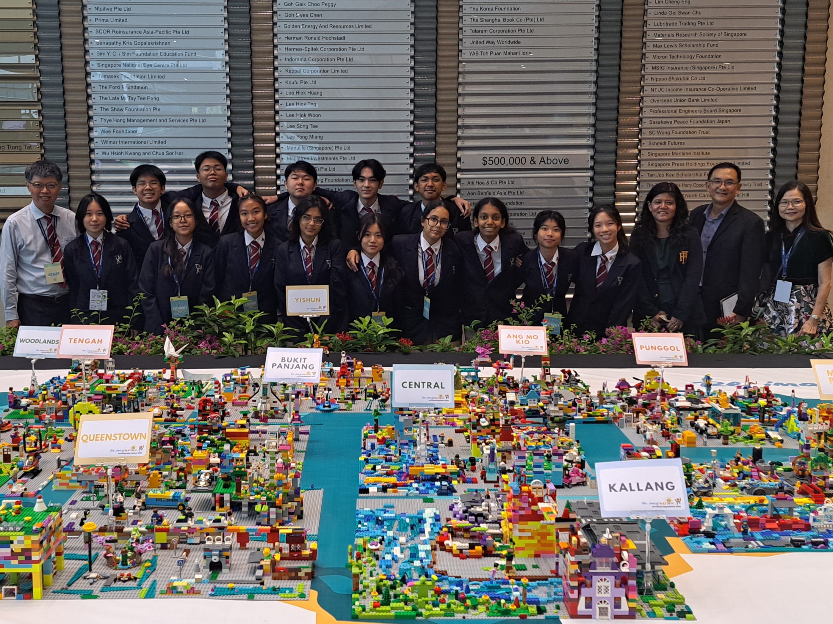 A group of students and teachers in school blazers posing behind a large colourful LEGO model of Singapore, with district labels such as Kallang, Queenstown, and Yishun visible.